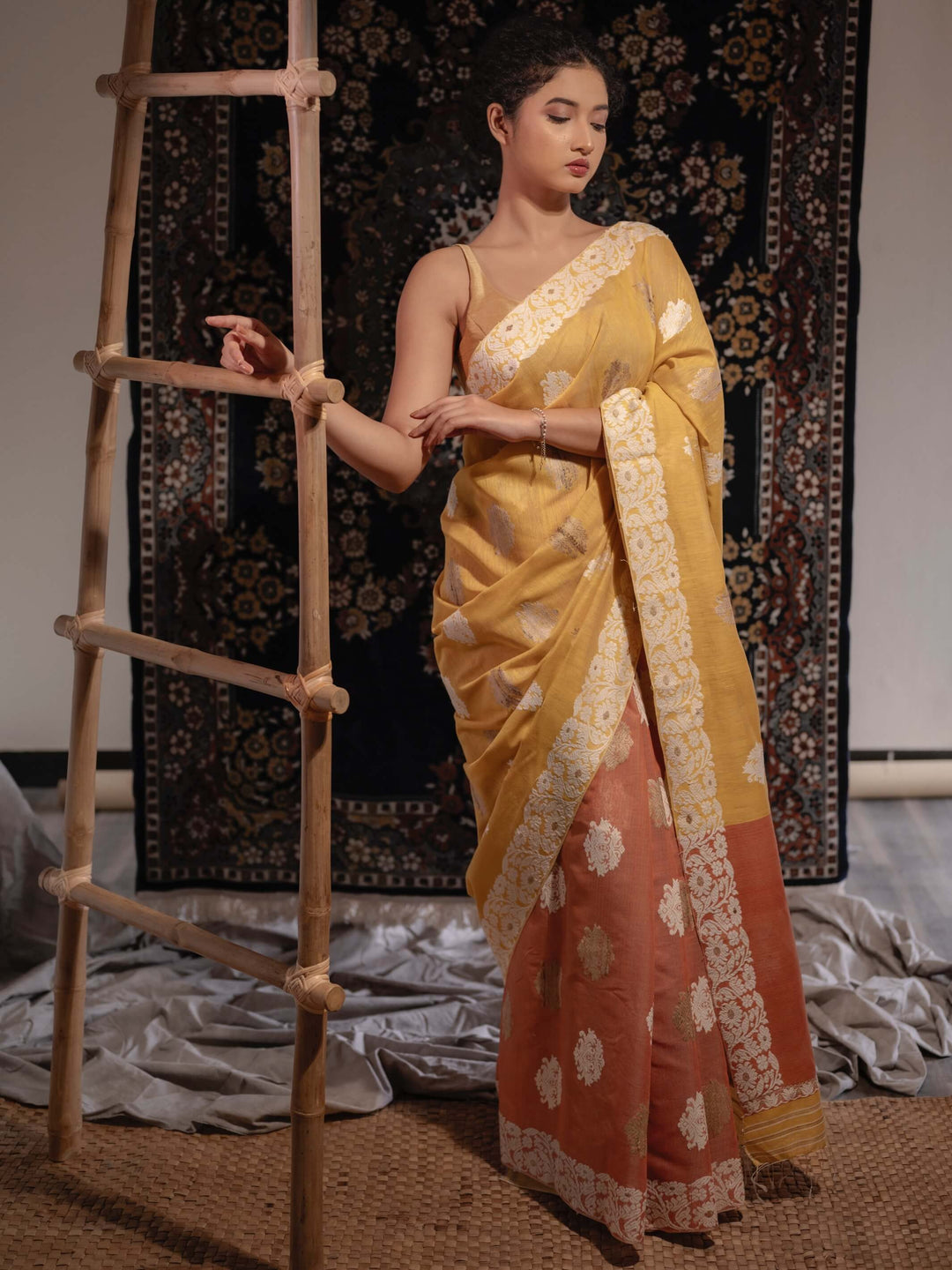 A model showcasing a naturally-dyed mustard yellow and lac rust orange silk saree with floral and paisley motifs and a woven floral border.