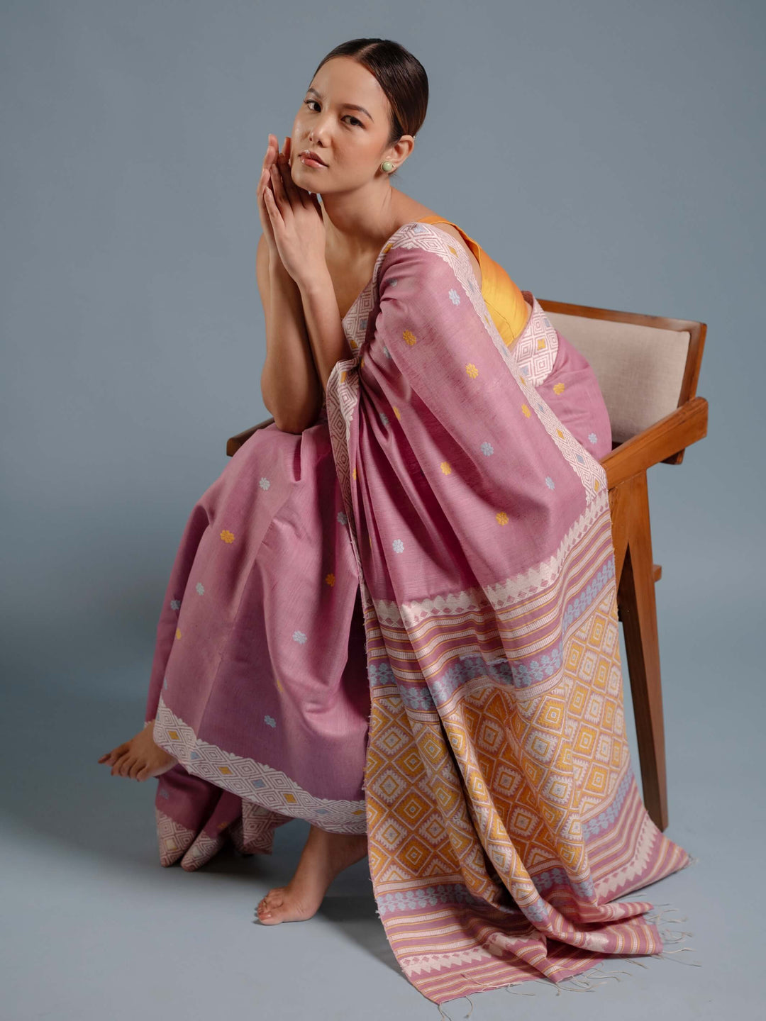 Woman sitting on a chair wearing a lilac pink saree with a patterned design against a gray background