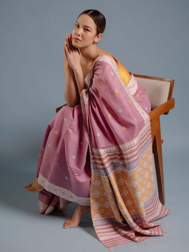Woman sitting on a chair wearing a lilac pink saree with a patterned design against a gray background