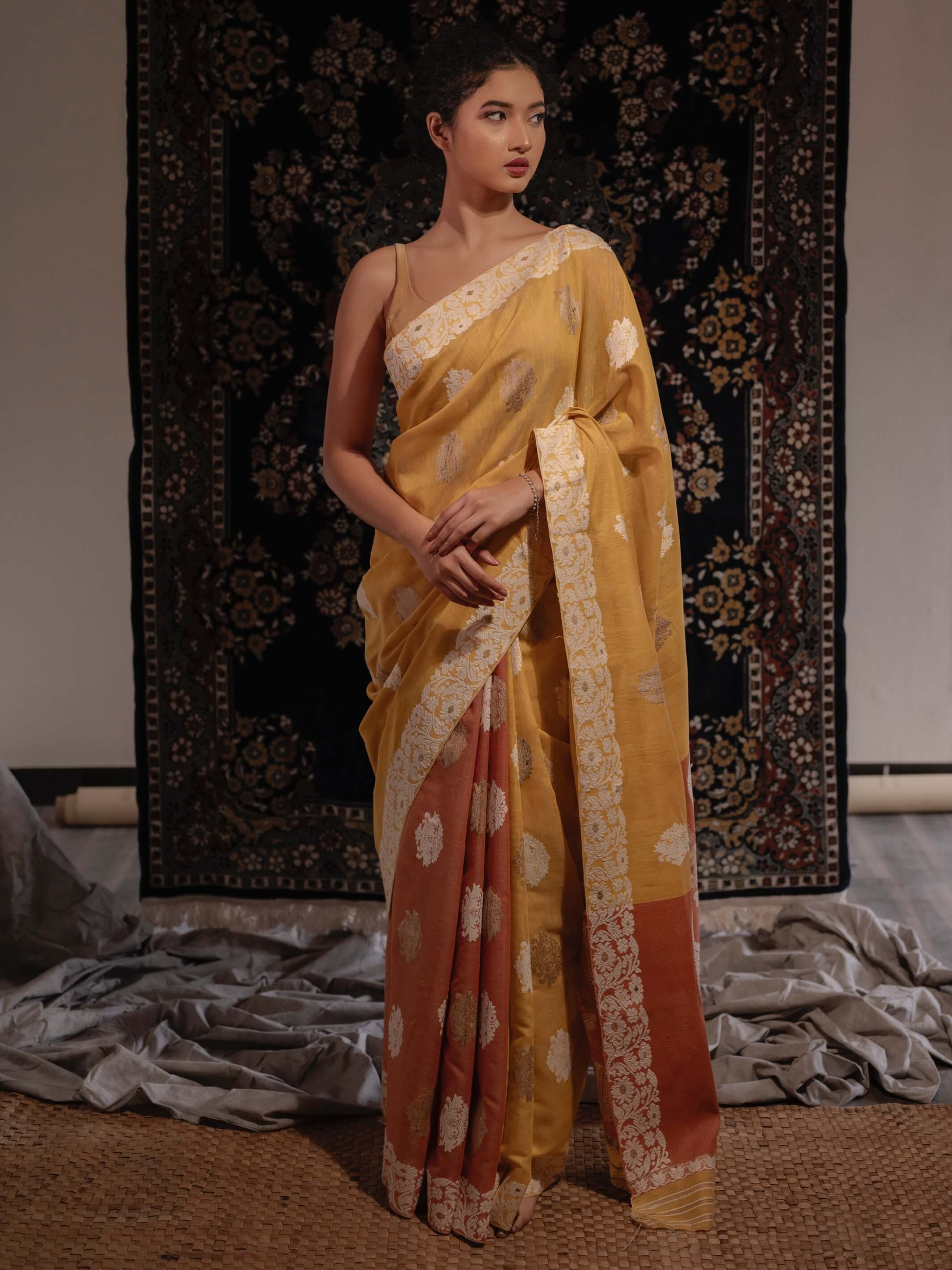 A model showcasing a naturally-dyed mustard yellow and lac rust orange silk saree with floral and paisley motifs and a woven floral border.