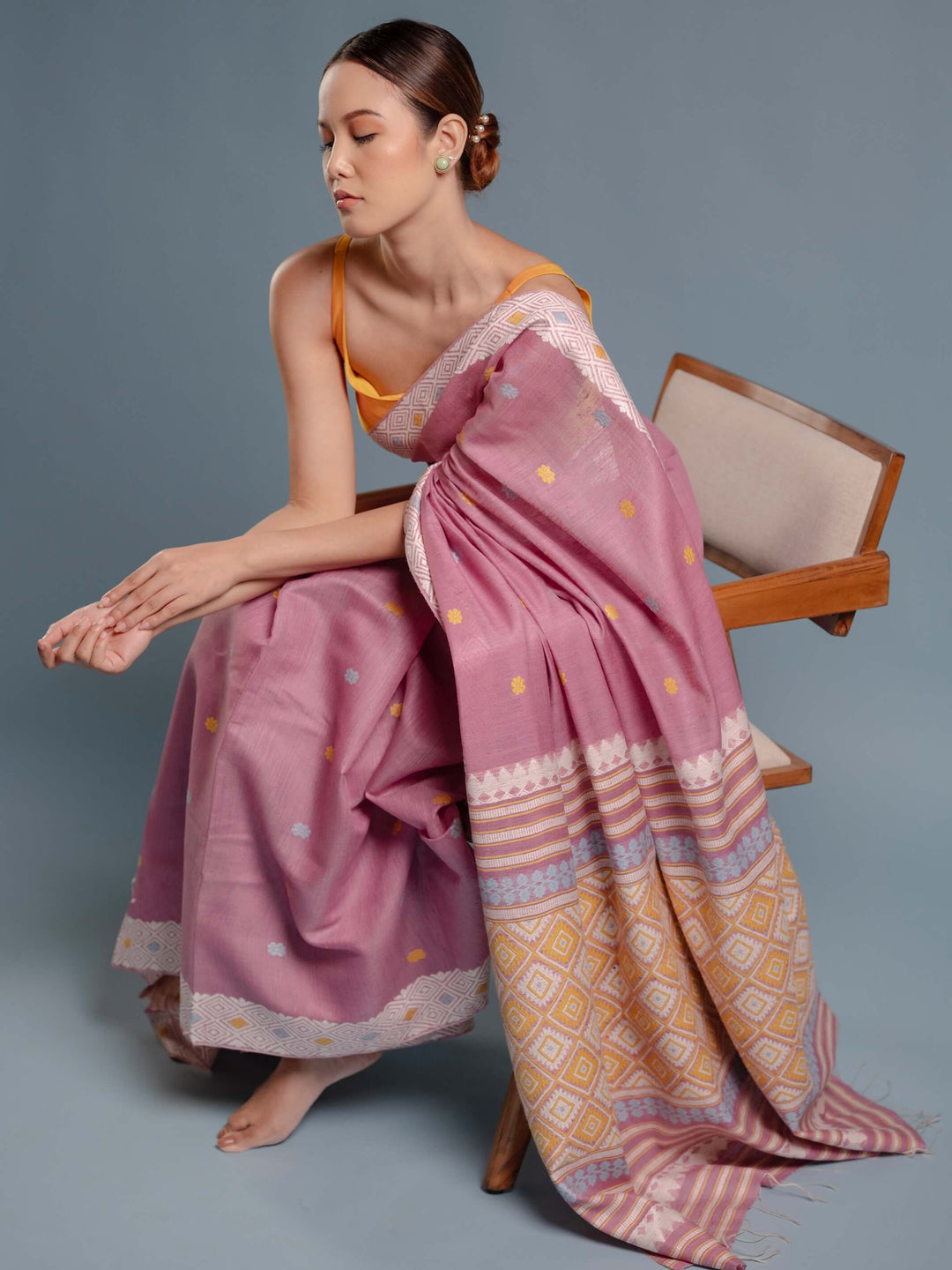 Woman wearing a lilac pink saree with a geometric pattern, sitting on a chair against a gray background