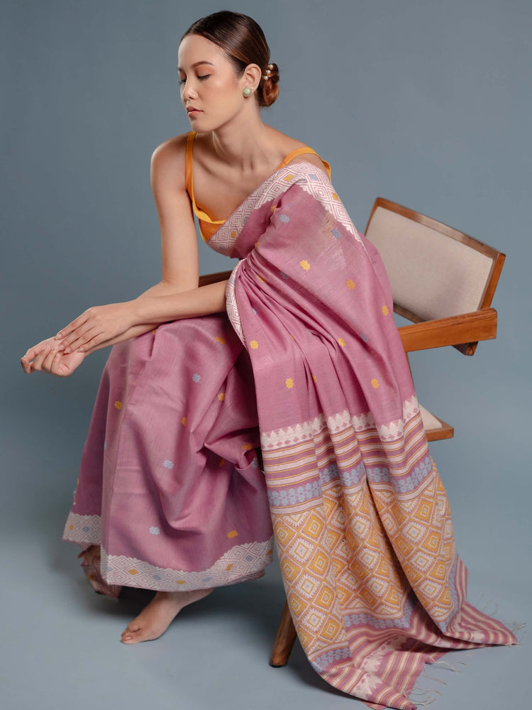 Woman wearing a lilac pink saree with a geometric pattern, sitting on a chair against a gray background