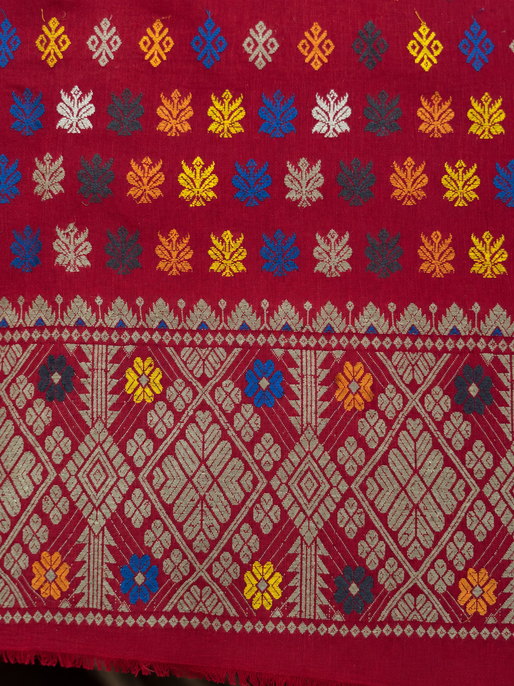 A close-up of a red woven textile with intricate traditional patterns, featuring rows of colorful floral motifs in yellow, blue, white, and gray, and a wide central band with geometric diamond and leaf designs.