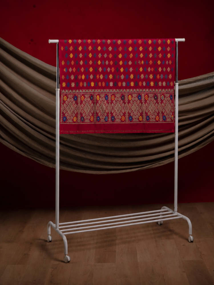 A red patterned textile hanging on a white metal rack against a dark red backdrop with draped brown fabric and a wood floor.