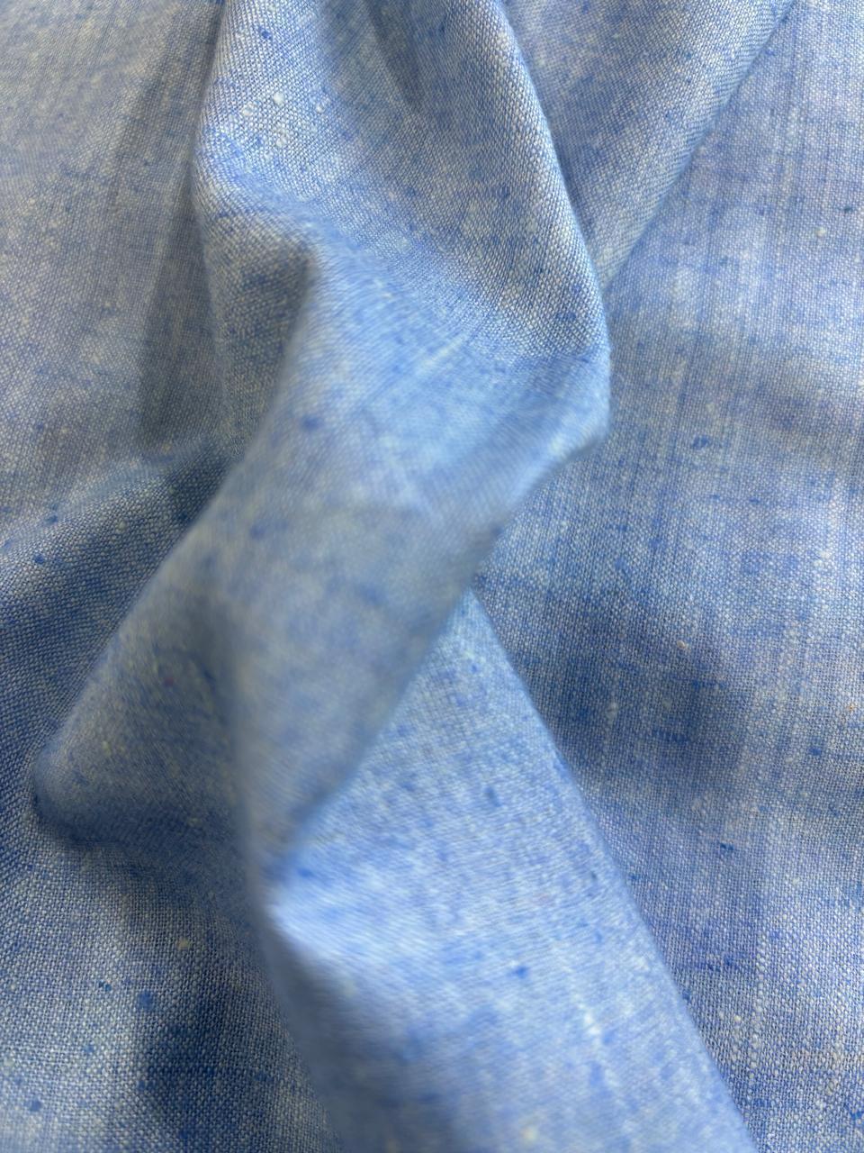 A close-up of light blue fabric with a subtle woven texture and small scattered blue flecks, draped in soft folds.