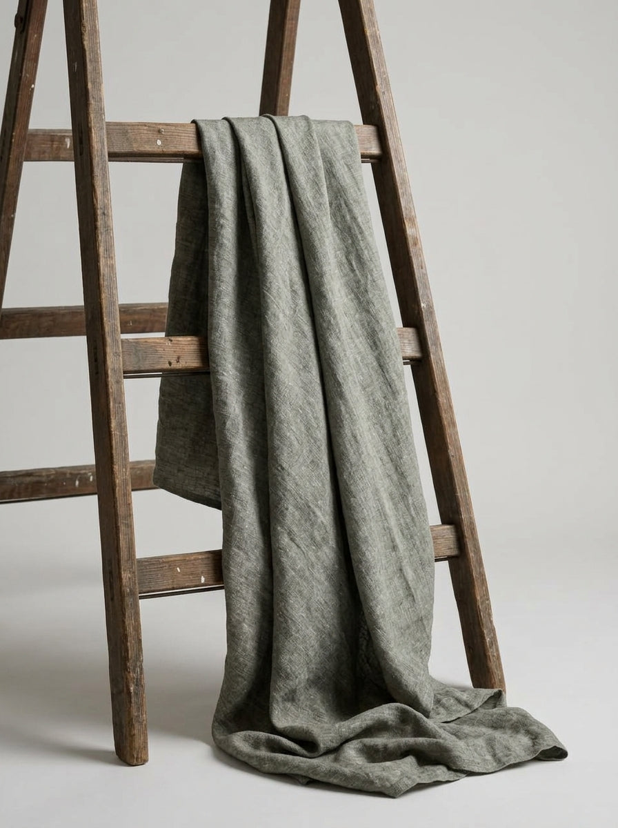 A textured, muted grey fabric draped over a rustic wooden ladder against a neutral light gray background.