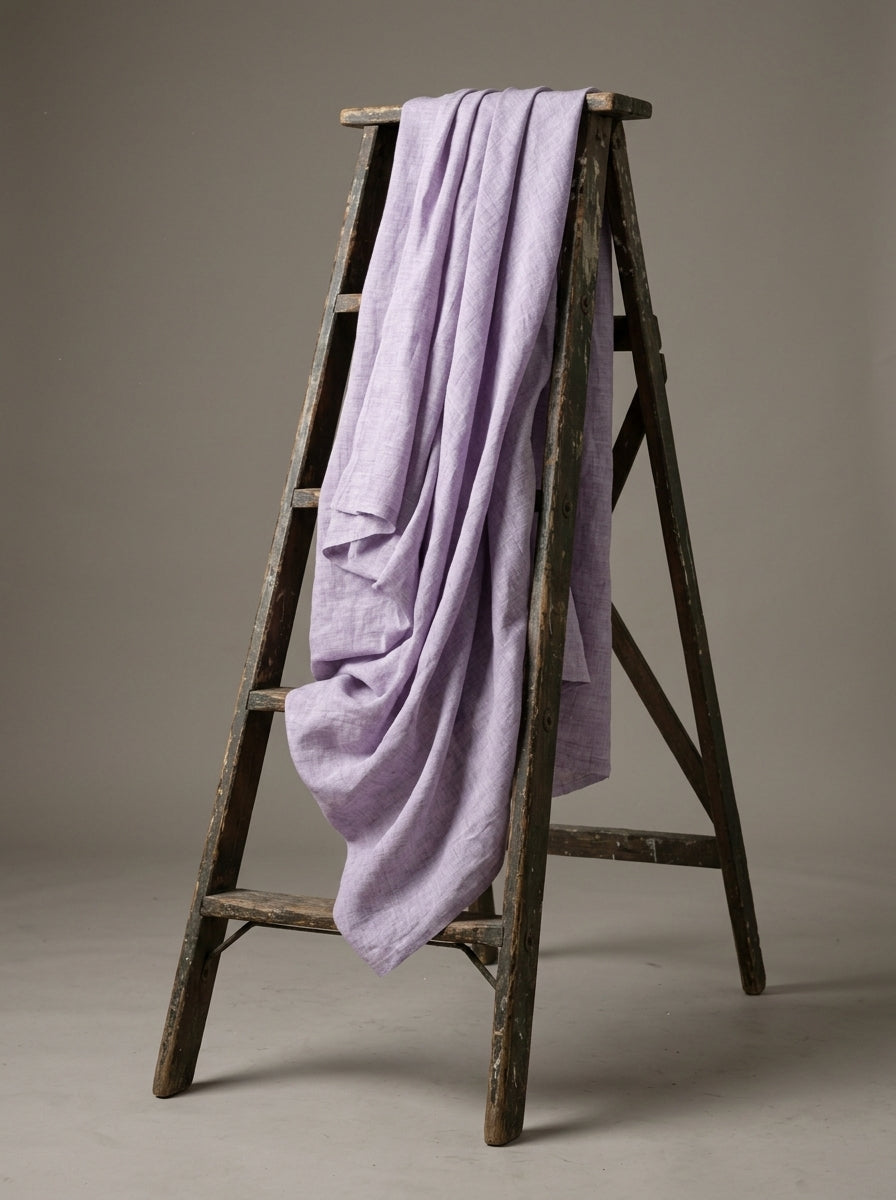 A light purple leri fabric draped over a rustic dark wooden A-frame ladder, photographed against a neutral warm gray studio background.