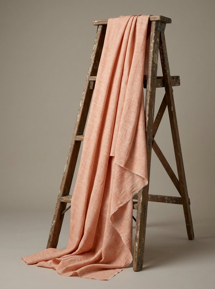 A peach-pink eri fabric draped over a rustic wooden A-frame ladder against a neutral taupe background.