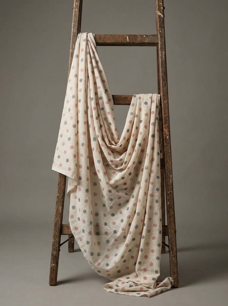 A cream-colored lightweight Eri silk fabric, with a pattern of small pink and gray polka dots, draped over a rustic wooden ladder against a neutral gray background.