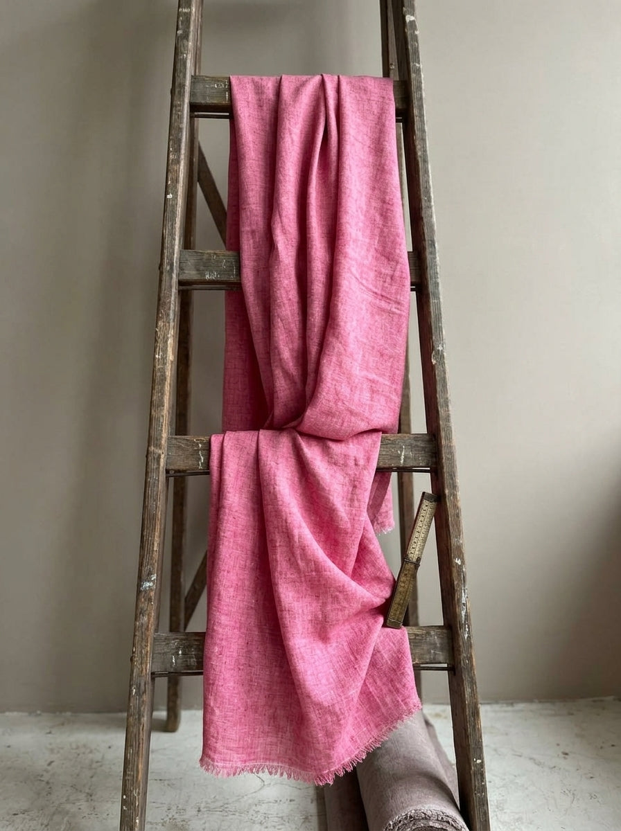 A pink eri handwoven fabric with fringed edges draped over a rustic wooden ladder against a neutral beige wall.